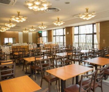 Ryokan dining room with wooden tables, chandeliers and large windows overlooking trees in Kusatsu, Gunma