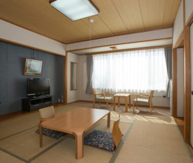 Spacious tatami room with low table, floor cushions, TV and seating by sunlit window at Itoen Hotel Kusatsu in Gunma