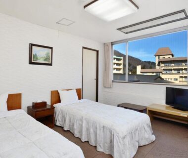 Bright twin guest room with two beds, large window overlooking town buildings and distant hills at Itoen Hotel New Sakura