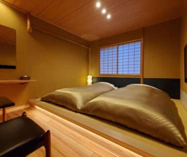 Traditional futon bedding on raised wooden platform, shoji screen window, dressing table and warm ambient lighting in ryokan room
