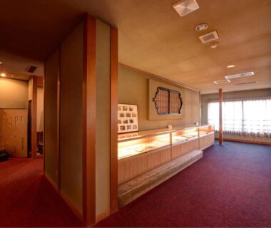 Carpeted ryokan corridor with glass display cases, wooden partitions, shoji window and warm ambient lighting at Iwamotoro in Kanagawa