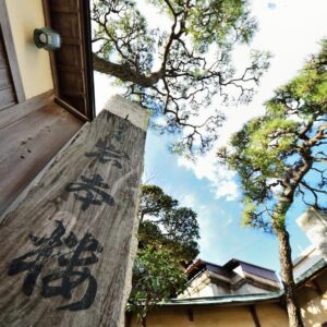 Wooden post inscribed with kanji at ryokan entrance, framed by pine trees and blue sky