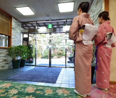 Two women in kimono standing in ryokan lobby by entrance overlooking a small garden and potted plants
