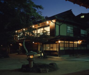 Traditional wooden ryokan exterior at night with lit shoji windows and stone lantern in courtyard