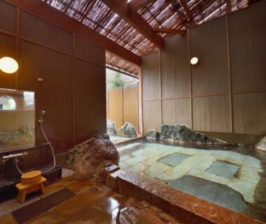 Indoor stone onsen bath with wooden slatted roof and shower area at Iyashi no Yado Iroha, Ōita