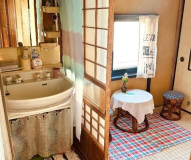Compact tatami room with shoji screens, low round table, patterned rug and adjacent washbasin area at Izu Hokkawa guesthouse, Shizuoka