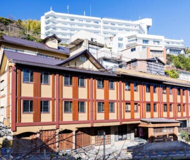 Traditional wooden ryokan facade with multiple stories on a hillside in Shizuoka, adjacent modern buildings