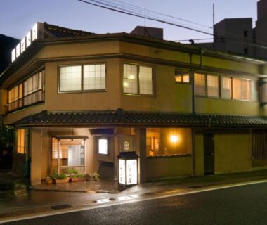 Lit exterior of two-story traditional ryokan with entrance lantern and tiled roof in Gifu