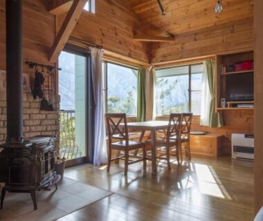 Sunlit wooden dining area with wood stove and large windows at Izumigo AMBIENT Azumino Cottage Nagano