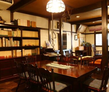 Warmly lit lounge with bookshelves, wooden table and chairs at Izumiya Zenbe in Nagano
