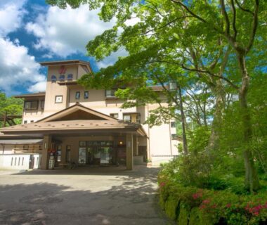 Front entrance of Jizaiso ryokan with tree lined grounds and paved driveway under blue sky