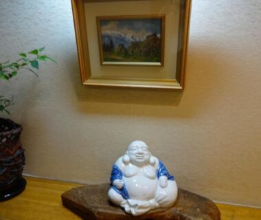 Smiling ceramic Buddha on wooden slab beneath framed painting and potted plant in Jizokan Matsuya, Nagano