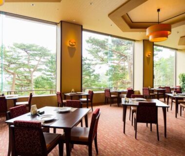 Spacious dining area with wooden tables, large windows and pine trees visible outside at Jodogahama Park Hotel Iwate