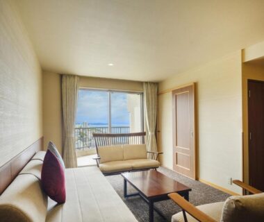 Bright seating area with sofa, bench and low table opening to balcony with distant sea view at KAMENOI HOTEL Atami