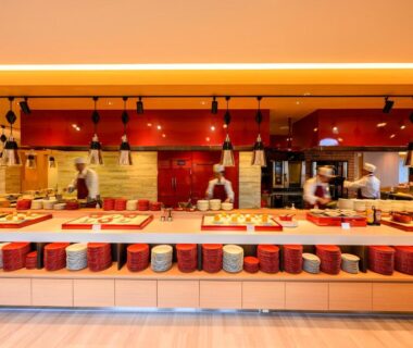 Open buffet counter with stacked bowls and chefs preparing dishes behind a red service counter