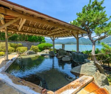 Outdoor stone onsen with wooden pergola overlooking coastal bay at KAMENOI HOTEL Nachi in Wakayama