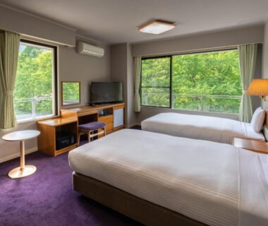 Bright twin room with large windows overlooking lush greenery, bedside lamps, desk, TV, seating at KAMENOI HOTEL Nikko Yunishigawa
