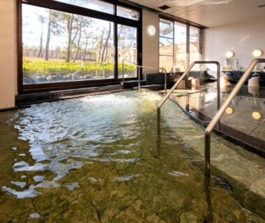 Spacious indoor onsen with stone floor, handrails and large windows facing a garden at KKR Numazu Hamayu, Shizuoka