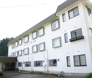 White three-story ryokan building with entrance canopy and forested hillside backdrop in Kansai