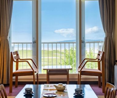 Tatami room with low table and chairs by sliding doors opening to balcony and green fields at Kagariya, Hokkaidō