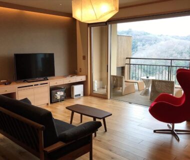 Wood-floored sitting area with sofa, red lounge chair, TV and balcony overlooking wooded hills at Kakimotoya Nara