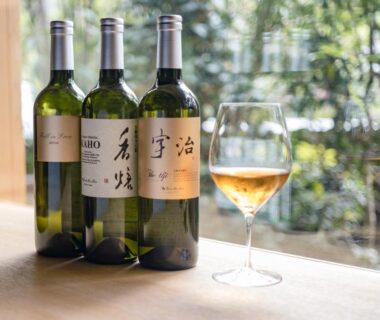 Three bottles with Japanese labels and a wine glass on a wooden counter by a window with garden view