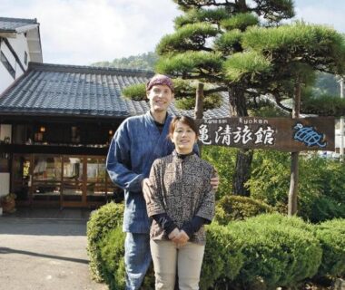 Two guests standing by Kamesei Ryokan entrance with pine tree and signboard in Nagano