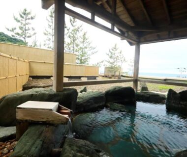 Covered rock-lined outdoor onsen with water view and wooden roof at Kameya Hotel in Yamagata