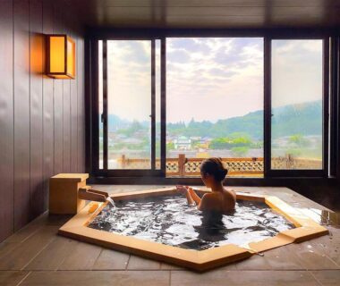 Guest soaking in a wooden indoor onsen tub with wide windows overlooking Chiba countryside