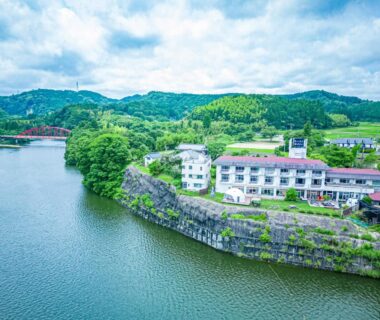 Lakeside building on rocky cliff at Kameyama Onsen Hotel surrounded by forested hills in Chiba, Kantō