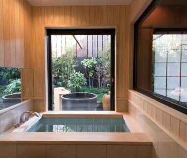 Indoor wooden soaking tub with outdoor round wooden tubs visible through garden window at Kamishichiken Oku Kyoto