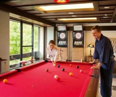 Two guests in robes play billiards on a red table beside large windows with garden view at Kanazawa Hyakurakusou Ishikawa