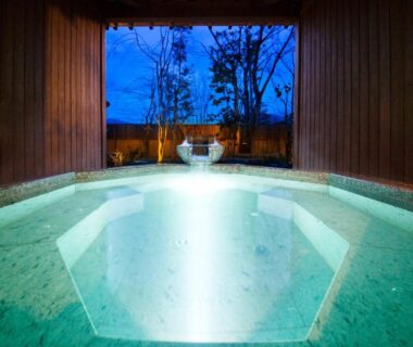 Illuminated indoor onsen bath with wooden interior and open view to trees at Kanazawa Hyakurakusou, Ishikawa