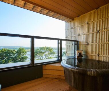 Circular wooden bath on balcony with bamboo wall at Kangetsuen in Hokkaidō overlooking water and trees