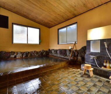 Stone-lined indoor onsen with tiled floor, wooden ceiling, shower stools and toiletries at Kannawa Yunoka in Ōita
