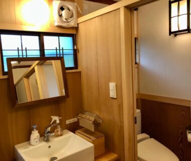 Compact wooden washroom with sink, angled mirror, and toilet at Kappo Ryokan Uoichi in Shizuoka