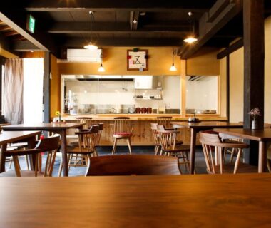 Wooden communal dining tables and chairs with open kitchen counter at Kariya Ryokan Q in Hyōgo, Japan