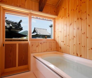 Wood-paneled bathing room with built-in tub and window overlooking tiled rooftops at Kariya Ryokan Q, Hyōgo