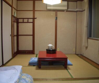 Tatami flooring with low red table, floor cushions, folded futon and wooden clothes rack at Kasuga Ryokan