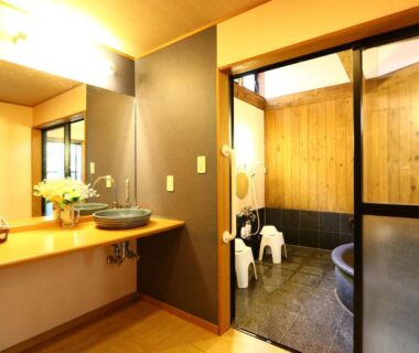 Wash basin and dressing area with sliding door leading to wood-paneled bath and stools at Kawachiya Ryokan, Mie