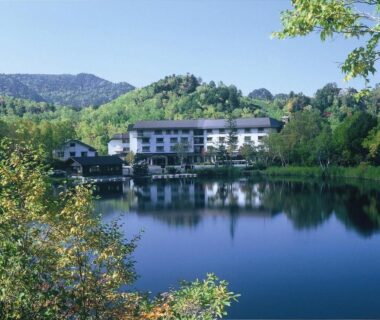 Lakeside hotel building reflected in calm lake surrounded by trees and forested hills at Kidoike Onsen Hotel Nagano