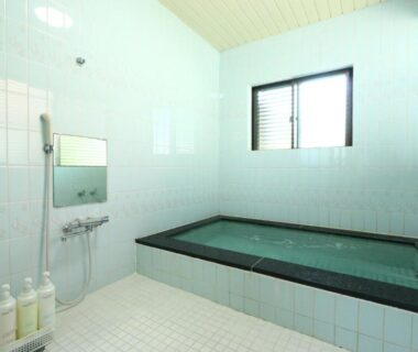 Rectangular tiled indoor bath with wall mounted shower, window and toiletries at a ryokan in Mie, Kansai
