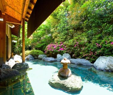 Stone-lined outdoor onsen with lantern, wooden eaves and flowering shrubs at Kikori, Yamanashi