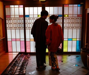 Two guests in kimono by stained glass shoji at ryokan entrance with wooden flooring and tubs