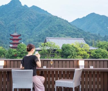 Guest seated on ryokan terrace with chairs, wooden railing, distant pagoda and forested mountains in Hiroshima