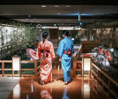 Two guests in yukata walking a lantern-lit wooden corridor with decorative umbrellas