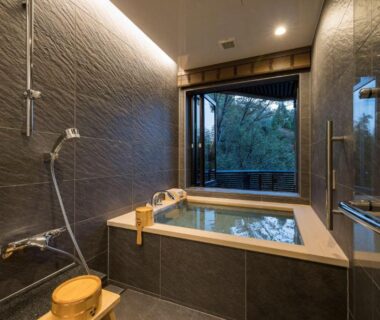 Indoor stone soaking tub with wooden bucket and shower, open window framing forest