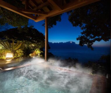 Steaming outdoor onsen bath under a covered veranda with lantern-lit garden and distant coastal waters at night