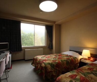 Twin guest room with floral bedcovers, bedside lamp, desk and window overlooking trees at Kitashiga Holiday Inn, Nagano