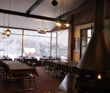 Ryokan dining area with central fireplace, wooden tables and chairs, large windows showing snow outside in Nagano